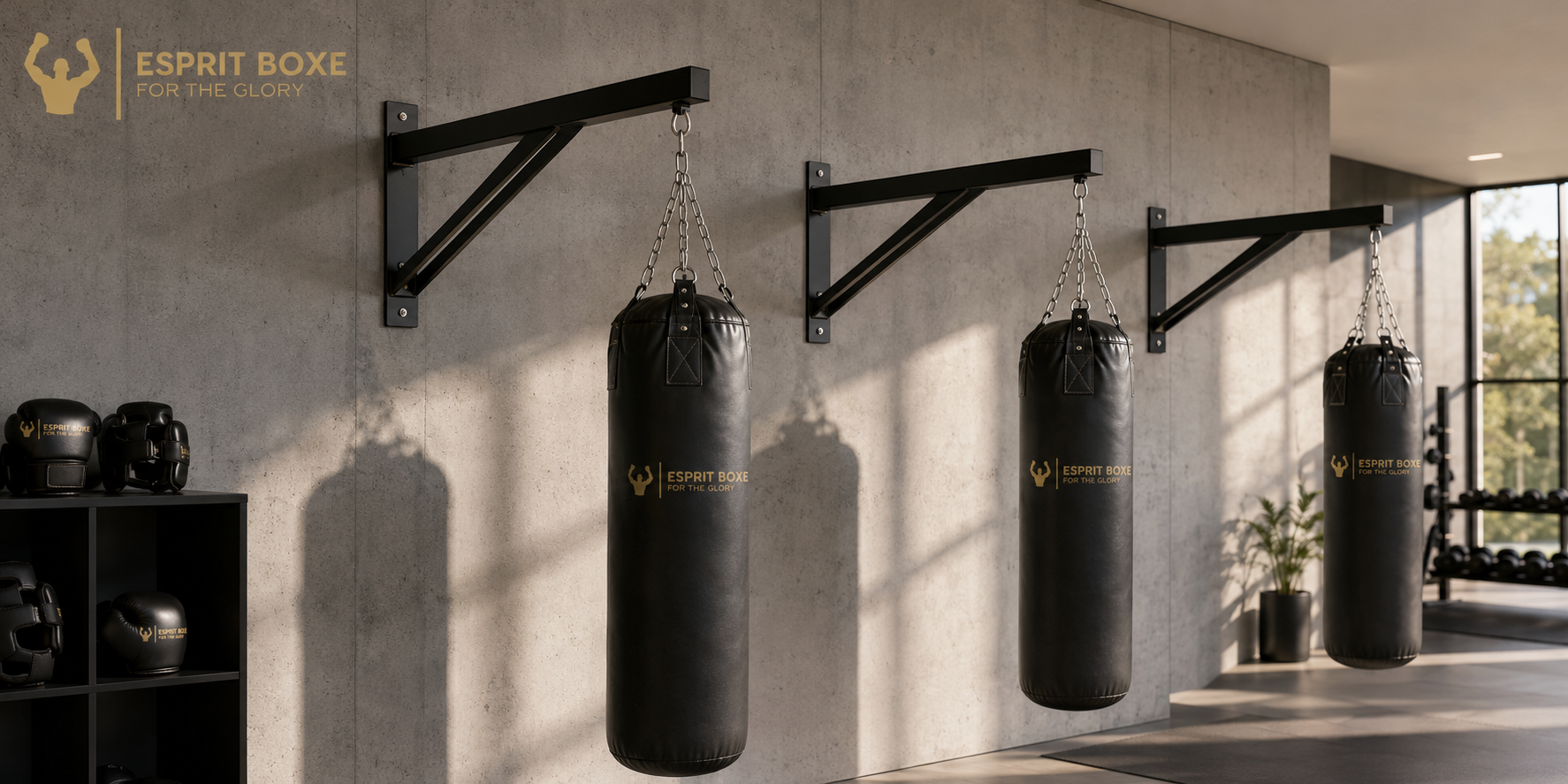 Collection potences pour sac de frappe Esprit Boxe : trois bras muraux en acier noir avec platine de fixation et jambe de renfort diagonale, chacun portant un sac de frappe noir à logo doré For the Glory suspendu par chaîne et mousquetons, sur un mur béton de salle