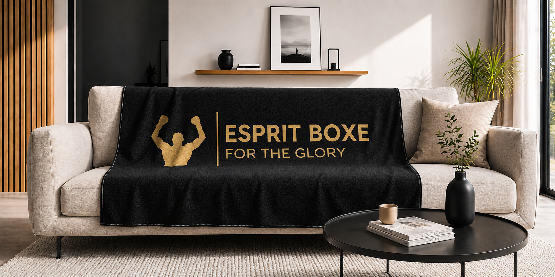 Collection plaids et serviettes de boxe Esprit Boxe : plaid noir a logo dore For the Glory drape sur un canape clair dans un salon